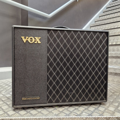 Pre-Owned Vox VT100X A Vox Valetcronix guitar amplifier speaker cabinet with a dark gray textured grille and a black diamond-patterned front, featuring gold 'VOX' lettering, placed on a metal diamond-plate surface with stairs in the background.