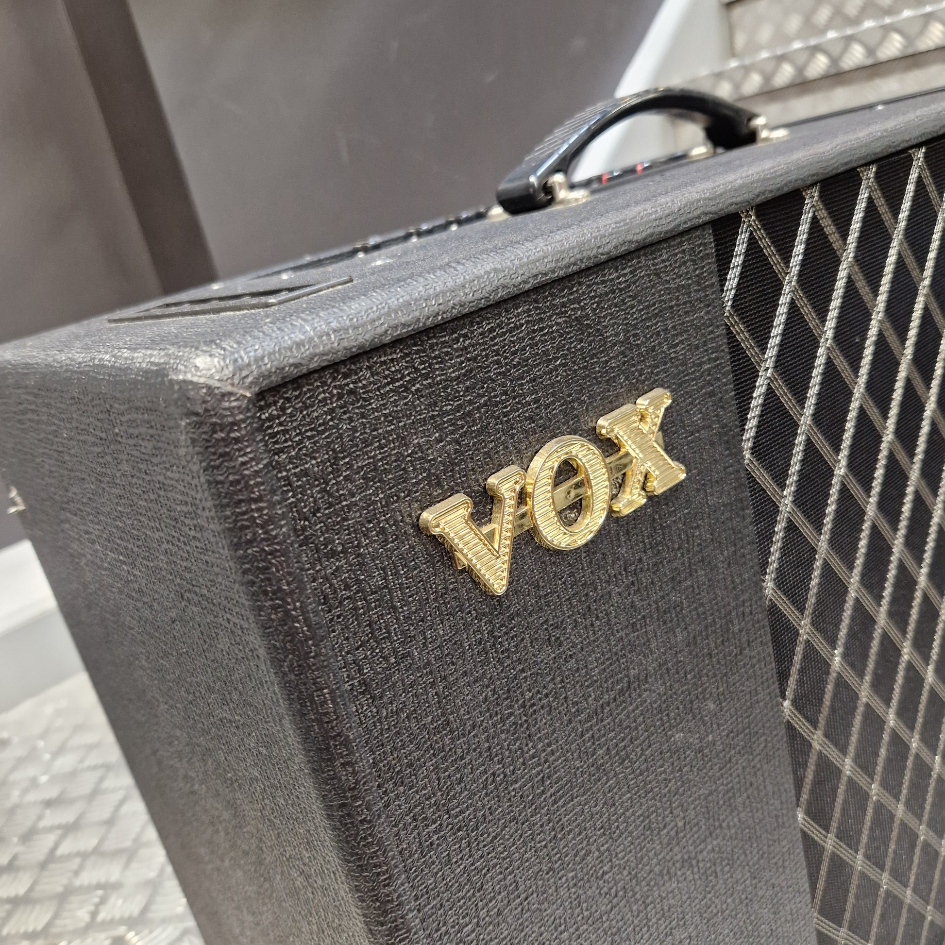 Pre-Owned Vox VT100X A gold-tone metal pin spelling 'MOT' with a textured, stone-like appearance, resting on the textured black fabric of a guitar amplifier's front panel.