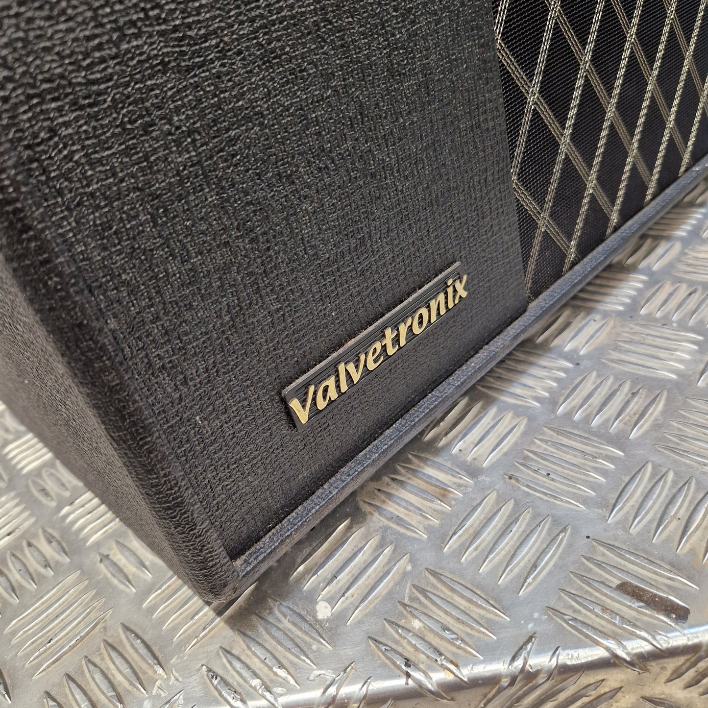 Pre-Owned Vox VT100X Close-up of a black textured Valvetronix speaker cabinet corner with a gold logo plate, resting on a silver diamond plate metal surface.