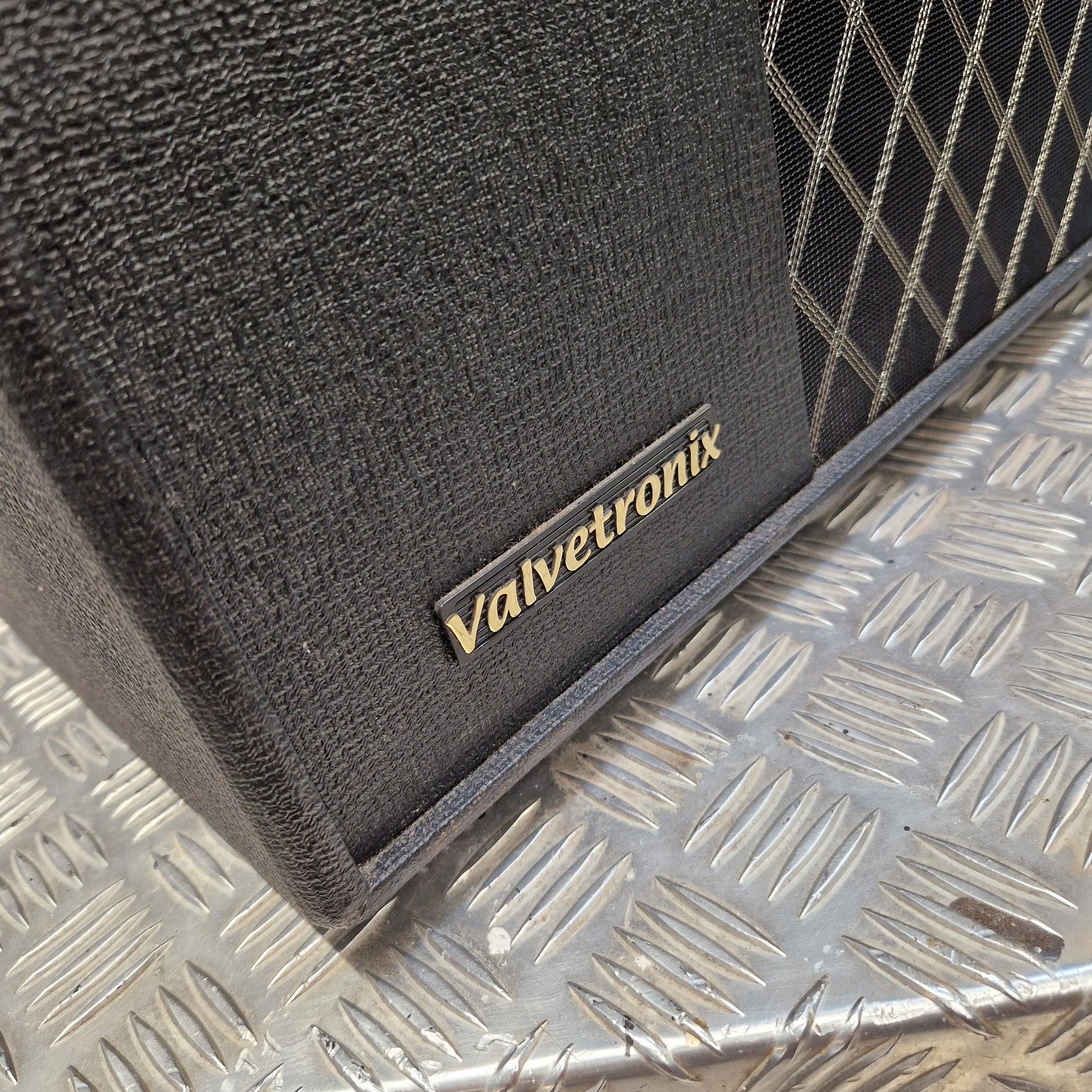 Pre-Owned Vox VT100X Close-up of a black textured Valvetronix speaker cabinet corner with a gold logo plate, resting on a silver diamond plate metal surface.