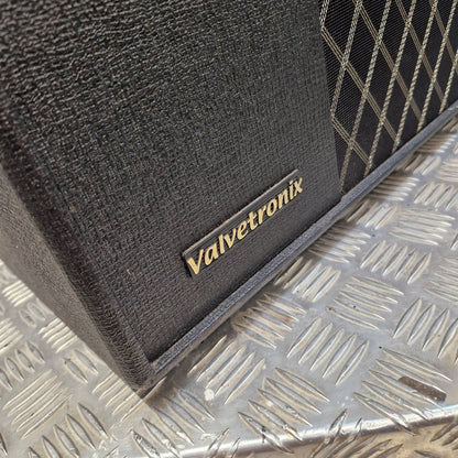 Pre-Owned Vox VT100X Close-up of a black textured Valvetronix speaker cabinet corner with a gold logo plate, resting on a silver diamond plate metal surface.