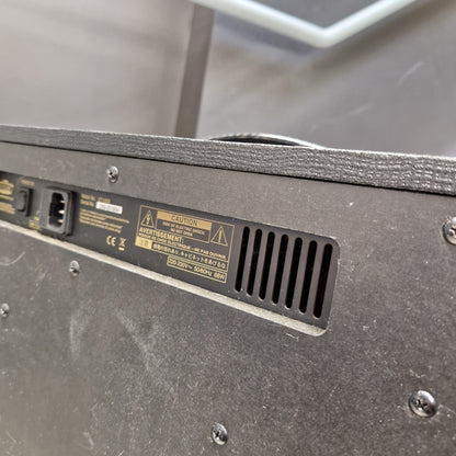 Pre-Owned Vox VT100X Close-up of the rear panel of a dark gray electronic equipment unit, showing a power inlet, ventilation grille, and printed warning labels.