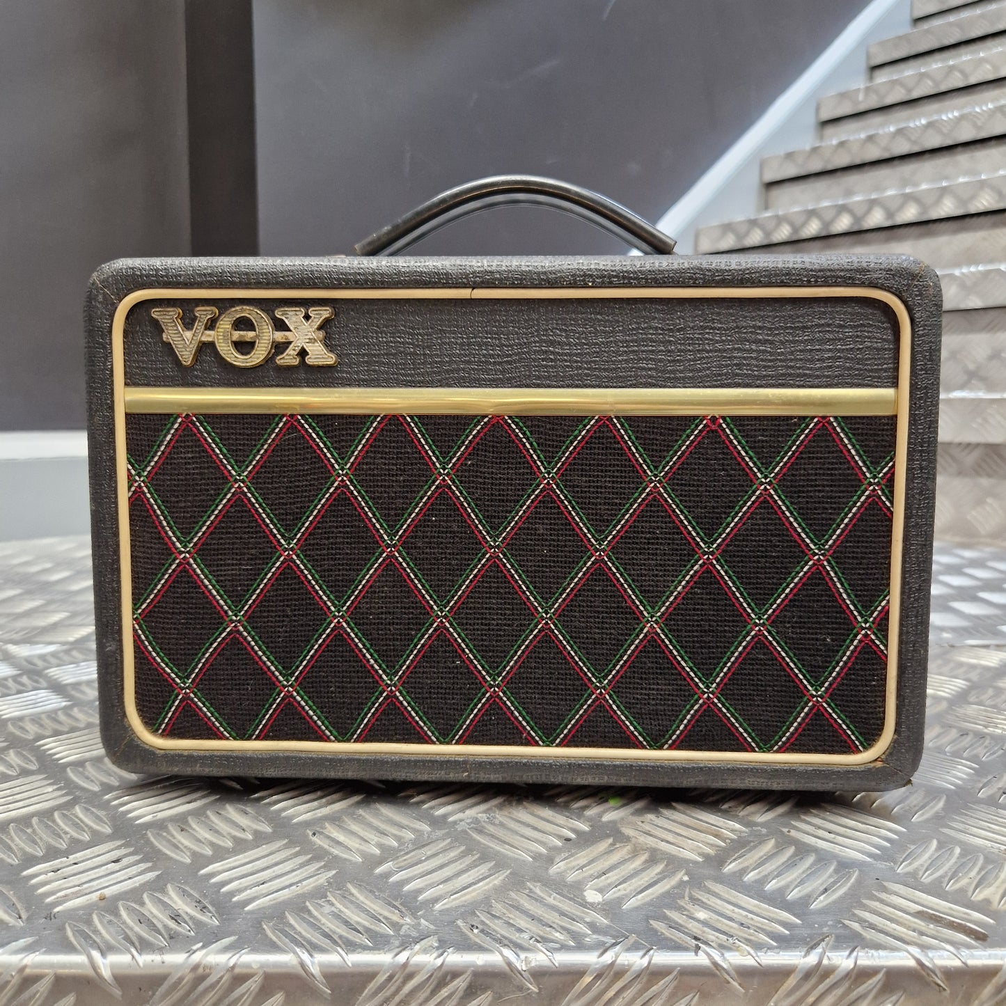 Pre-Owned Vox Escort A Vox portable speaker with a textured dark gray fabric cover, featuring a black diamond-patterned grille with red and green stitching, gold trim, and the gold Vox logo.
