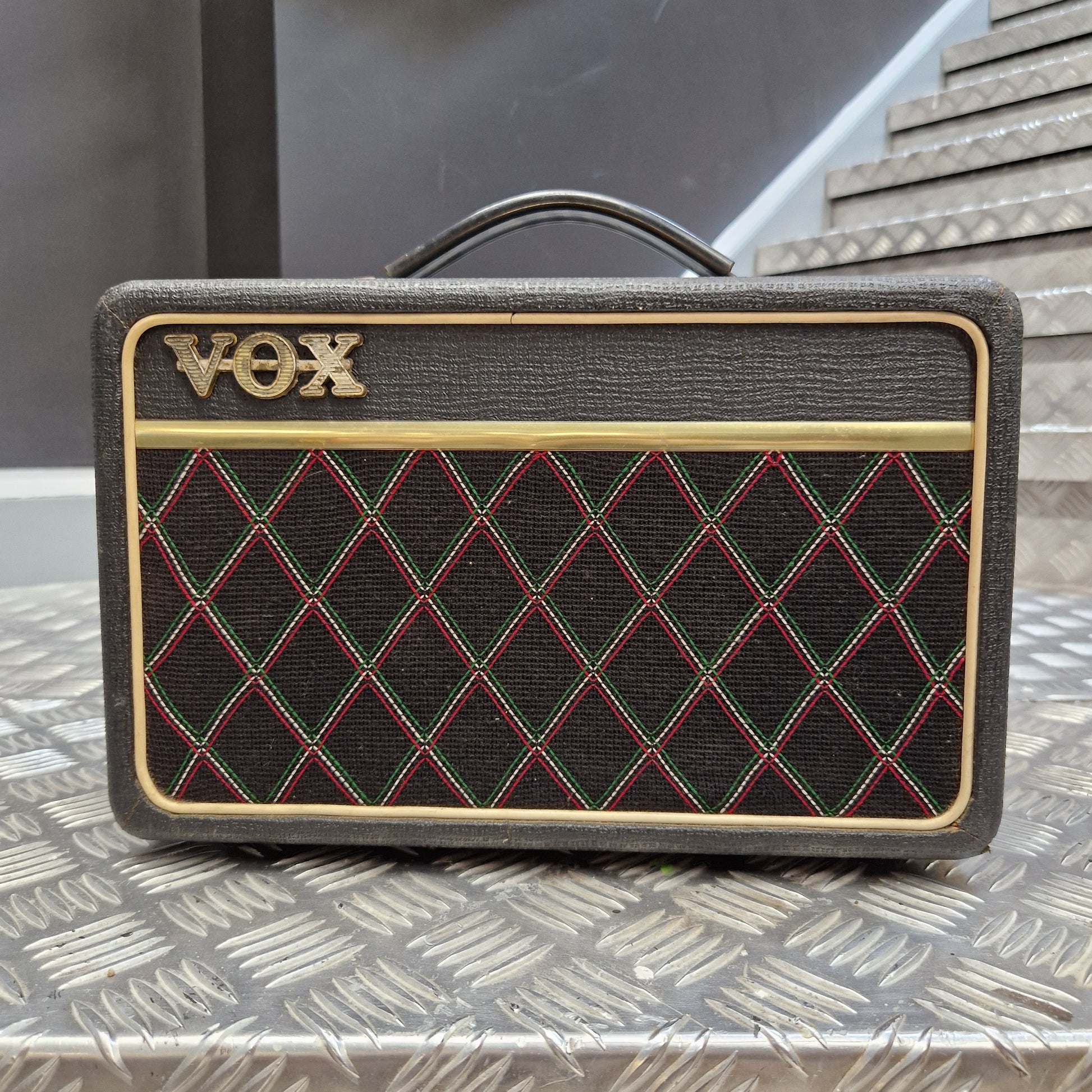 Pre-Owned Vox Escort A Vox portable speaker with a textured dark gray fabric cover, featuring a black diamond-patterned grille with red and green stitching, gold trim, and the gold Vox logo.