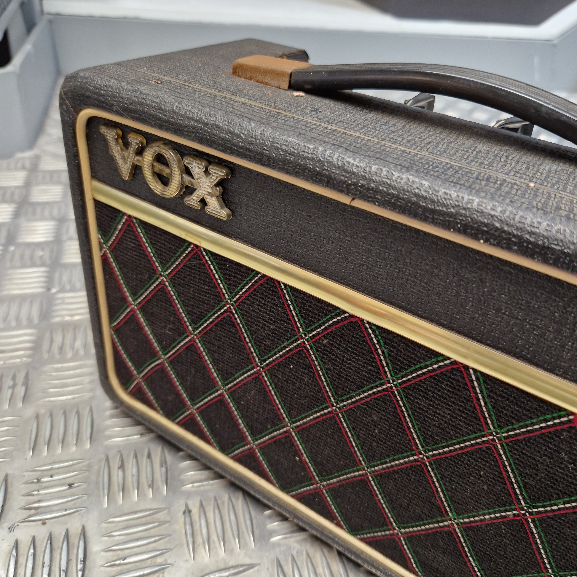 Pre-Owned Vox Escort A vintage Vox guitar amplifier head with a black textured grille cloth, gold trim, and red and green diamond-pattern stitching, resting on a silver diamond plate metal surface.