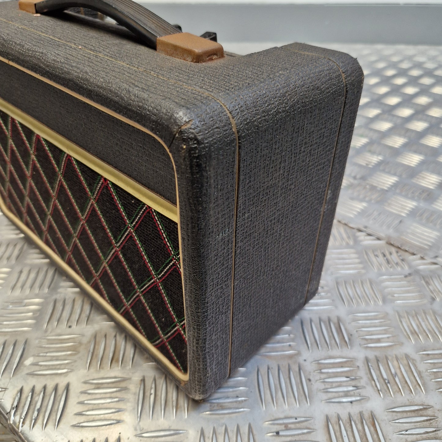 Pre-Owned Vox Escort A close-up of a vintage-style guitar amplifier or speaker cabinet with a dark gray textured fabric cover, tan trim, and a diamond-pattern speaker grille, resting on a metal diamond-plate surface.