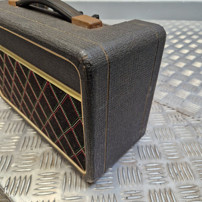 Pre-Owned Vox Escort A close-up of a vintage-style guitar amplifier or speaker cabinet with a dark gray textured fabric cover, tan trim, and a diamond-pattern speaker grille, resting on a metal diamond-plate surface.