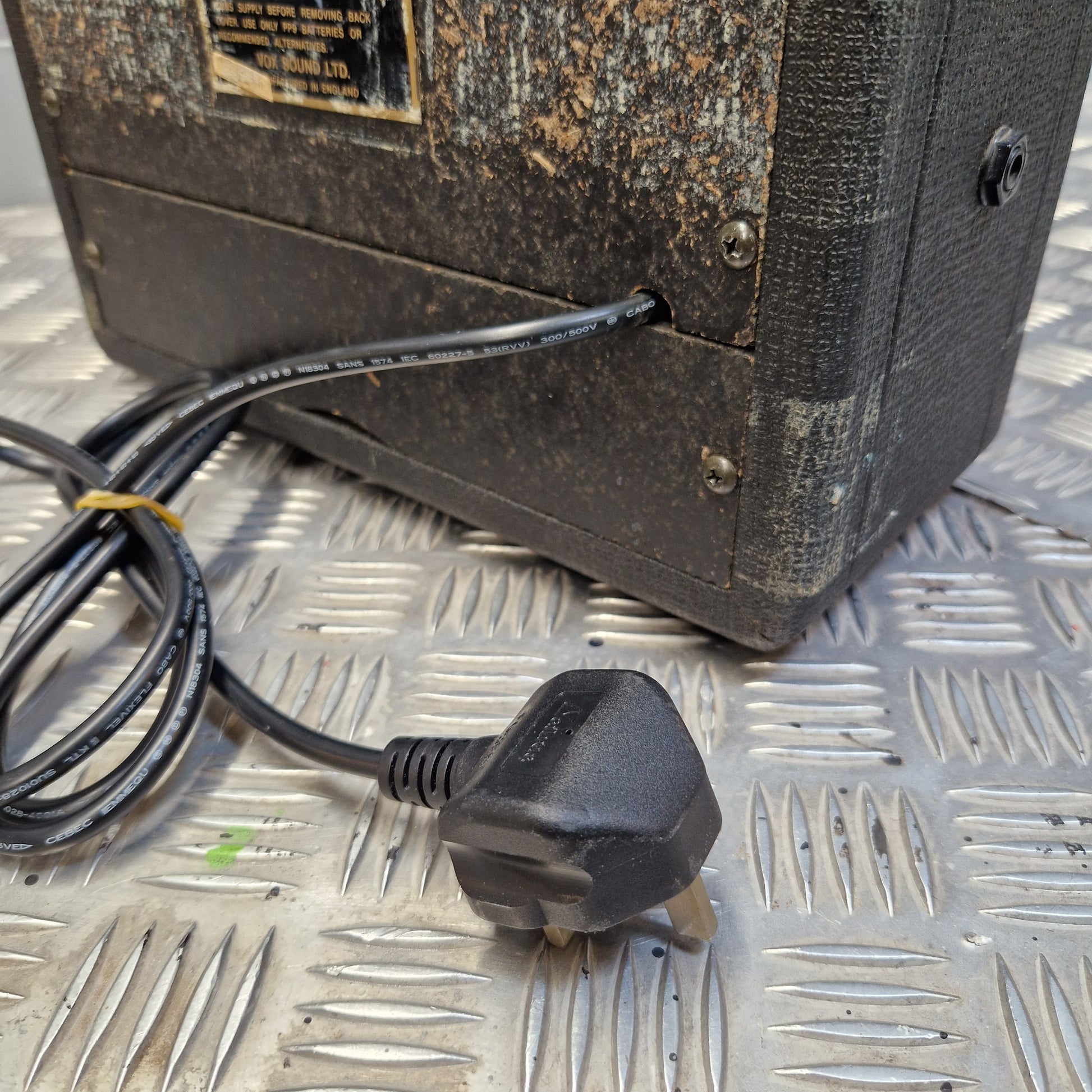 Pre-Owned Vox Escort A close-up of a black power cord with a UK three-pin plug resting on a silver diamond plate metal surface next to a weathered black equipment box.