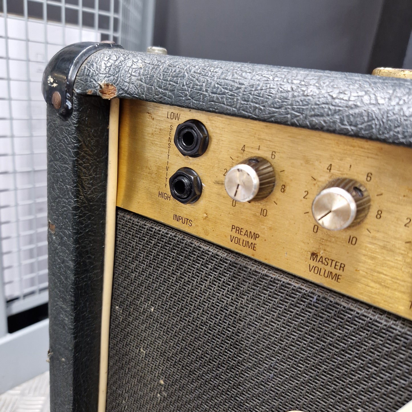 Pre-Owned Marshall JCM 800 Close-up of a vintage guitar amplifier corner showing a black textured vinyl cover, a gold-tone control panel with silver dials, and a black speaker grille.