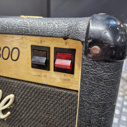 Pre-Owned Marshall JCM 800 Close-up of a vintage-style guitar amplifier corner with a black textured cover, gold-toned control panel, red power switch, and part of a white script logo on the speaker grille.