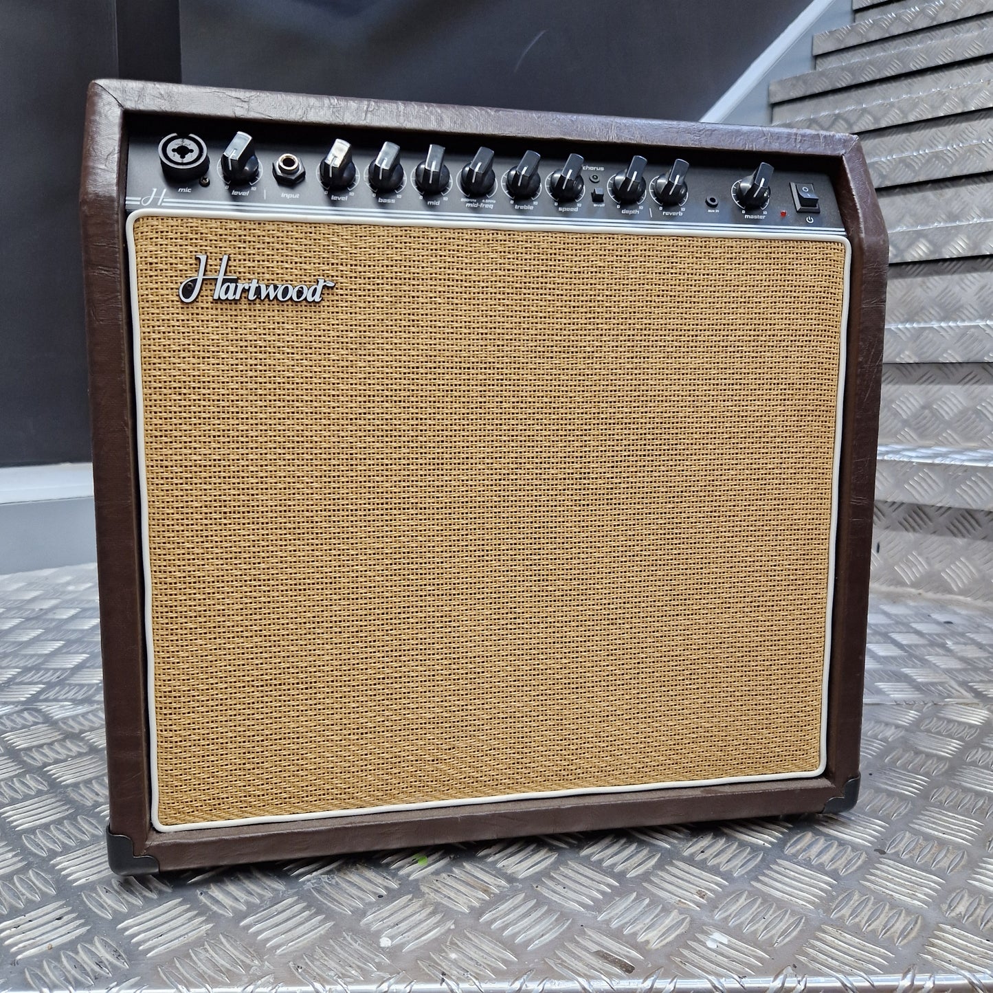Pre-Owned Hartwood HW-AA-30 A vintage-style Harman guitar amplifier with a brown wood-look cabinet, tan woven grille cloth, and black control knobs, photographed from above on a textured metal surface.