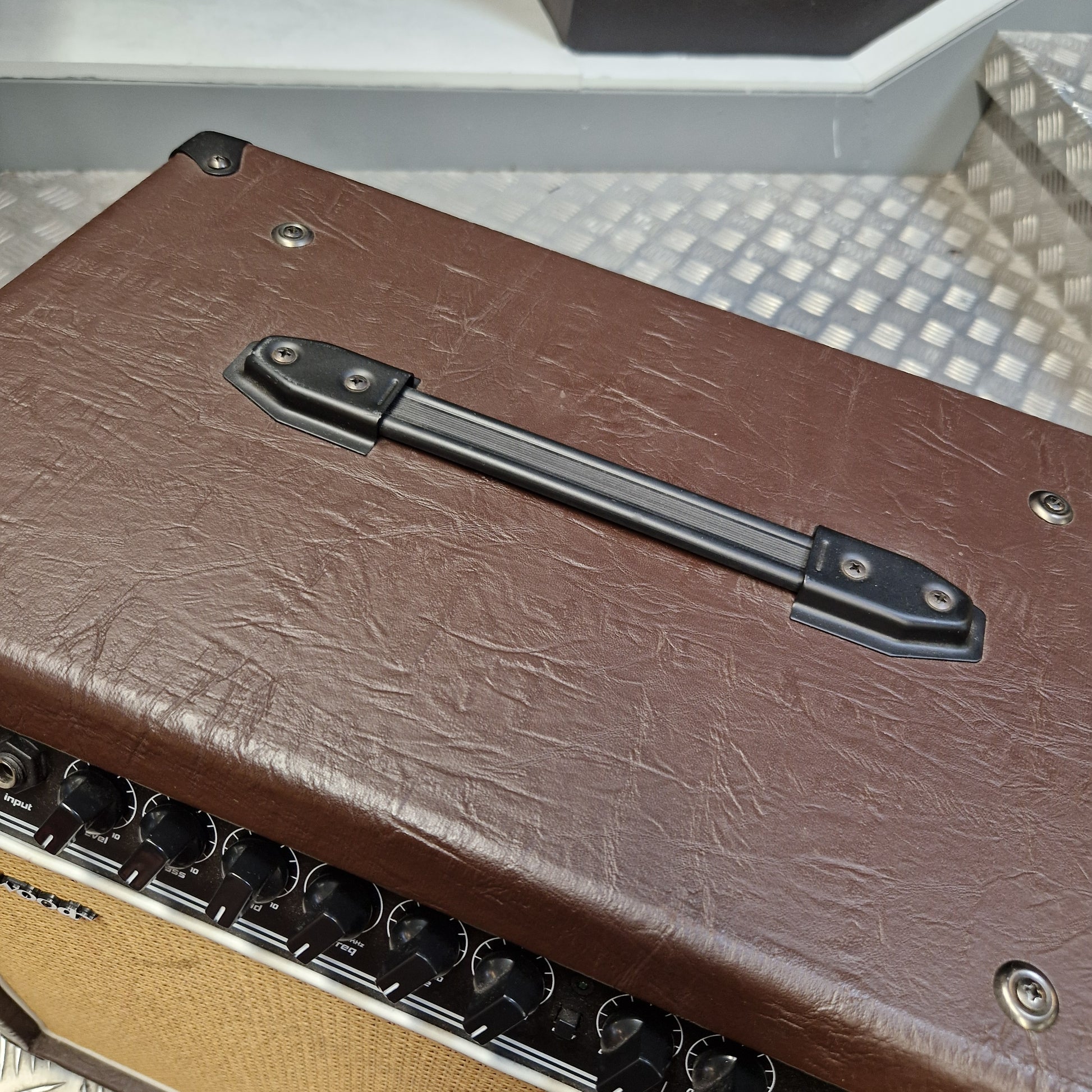 Pre-Owned Hartwood HW-AA-30 Close-up of the side of a brown textured amplifier or speaker cabinet with a black handle, metal screws, and part of the tan speaker grille visible.