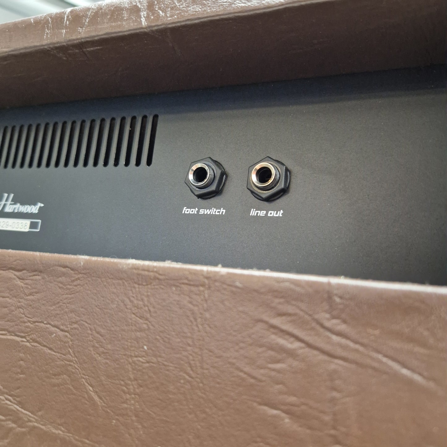 Pre-Owned Hartwood HW-AA-30 Close-up of the rear panel of a Harman speaker, showing two audio jacks labeled line out and hot switch, with a vented grille and partial Harman logo, framed by brown textured sides.