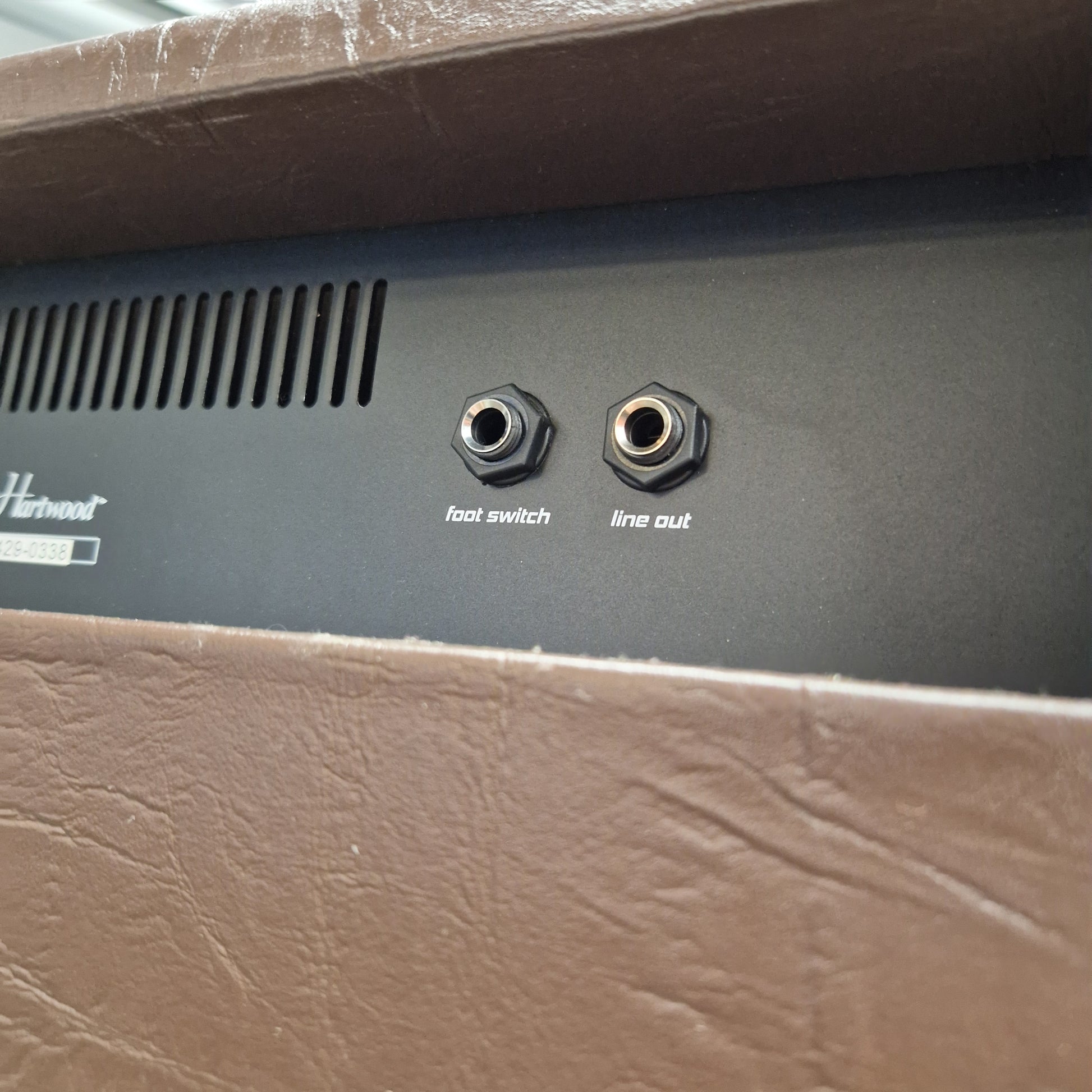 Pre-Owned Hartwood HW-AA-30 Close-up of the rear panel of a Harman speaker, showing two audio jacks labeled line out and hot switch, with a vented grille and partial Harman logo, framed by brown textured sides.