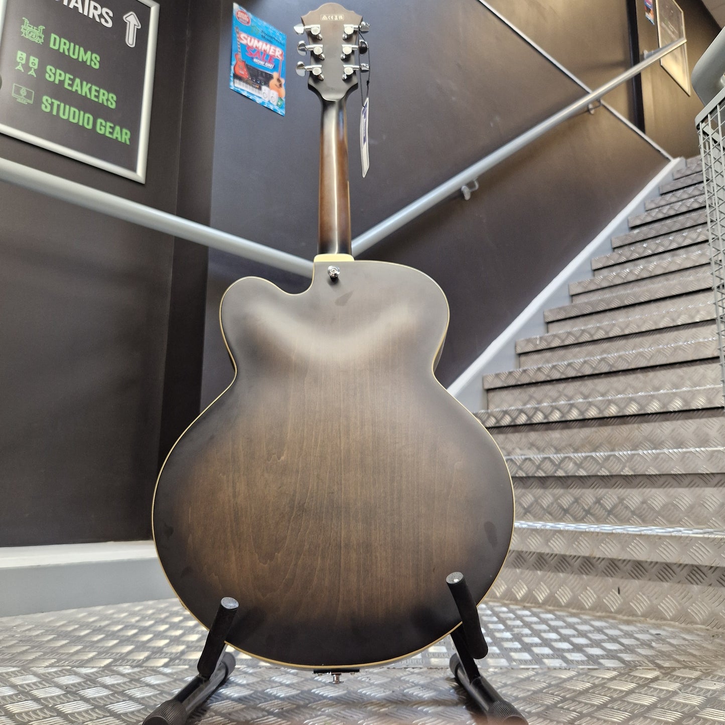 Pre-Owned Ibanez AF55-TKF Hollowbody Electric - Black Flat The back of an electric guitar with a dark brown sunburst finish, displayed on a stand in front of a staircase.
