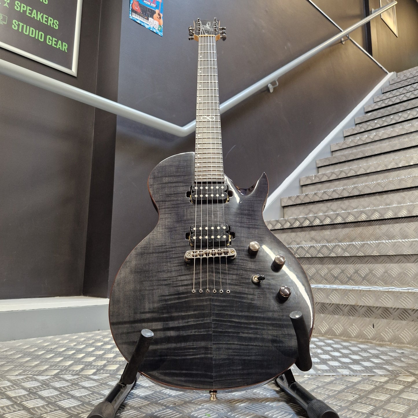 Pre-Owned Chapman ML-2 Electric - Grey A black electric guitar with a glossy flamed maple top and dark finish, resting on a stand in an indoor showroom with acoustic panels and stairs in the background.