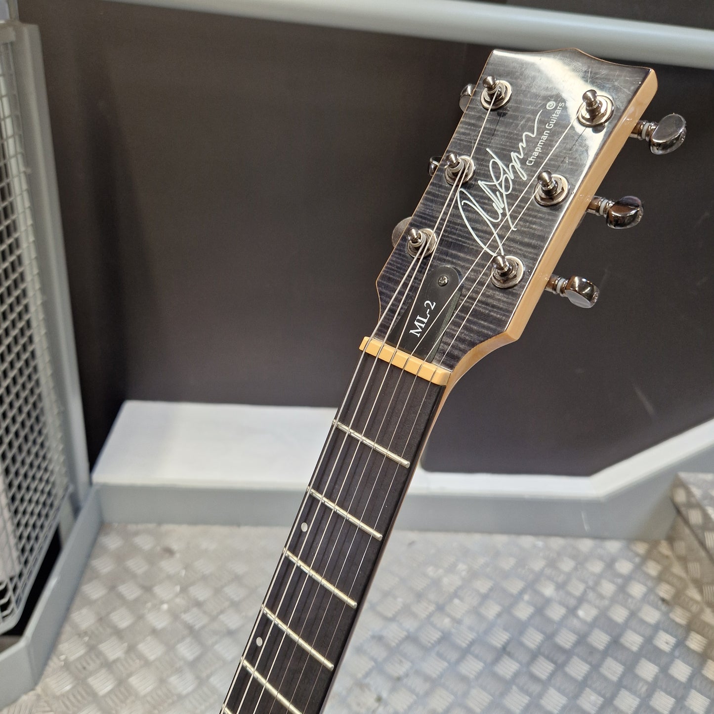 Pre-Owned Chapman ML-2 Electric - Grey Close-up of a guitar headstock and upper fretboard, showing the brand signature and model markings, photographed indoors against a dark and metallic background.