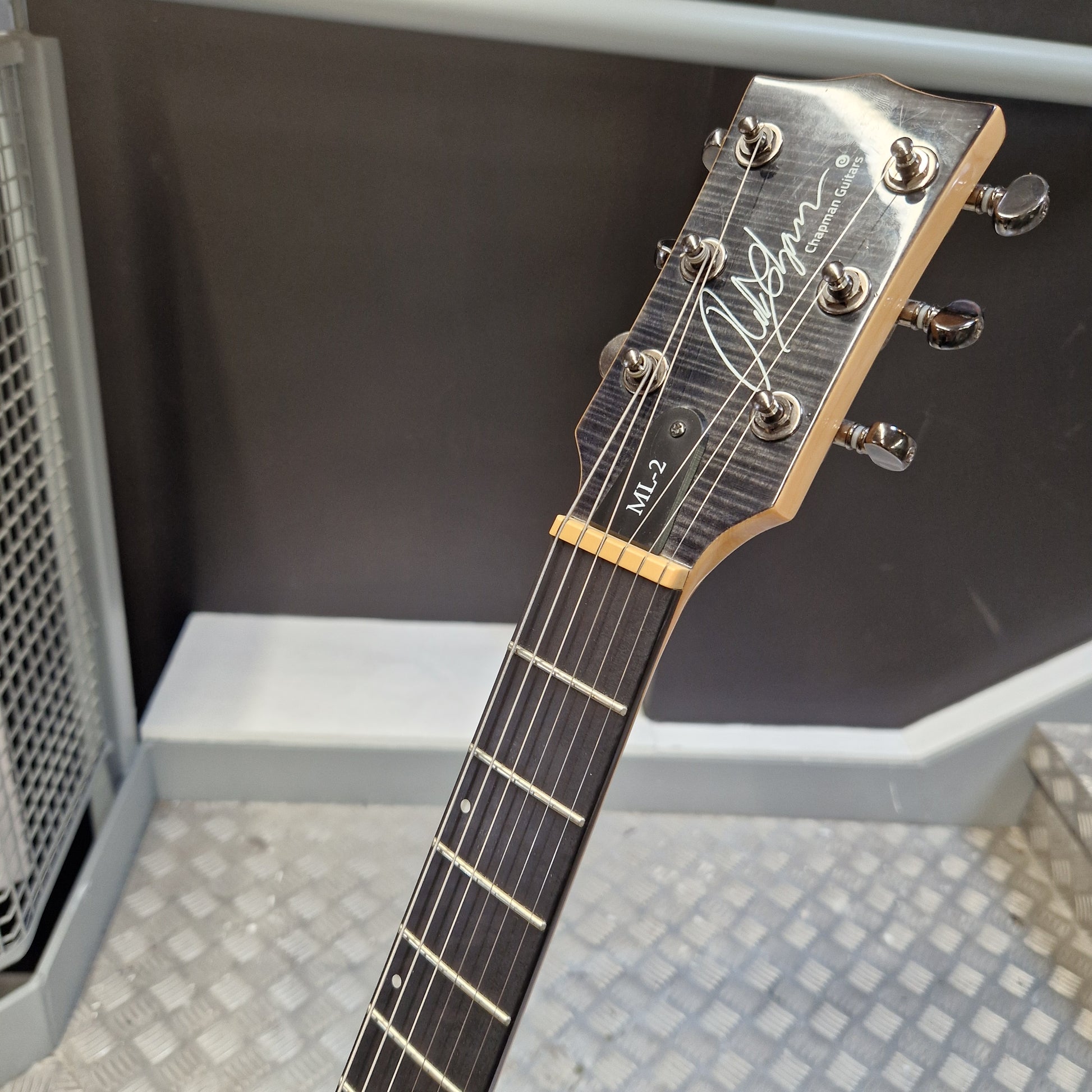 Pre-Owned Chapman ML-2 Electric - Grey Close-up of a guitar headstock and upper fretboard, showing the brand signature and model markings, photographed indoors against a dark and metallic background.