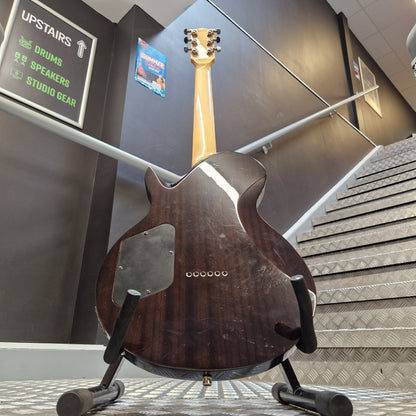 Pre-Owned Chapman ML-2 Electric - Grey A dark brown electric guitar body on a display stand inside a showroom, with part of the light wood neck visible and a metal diamond-plate wall in the background.