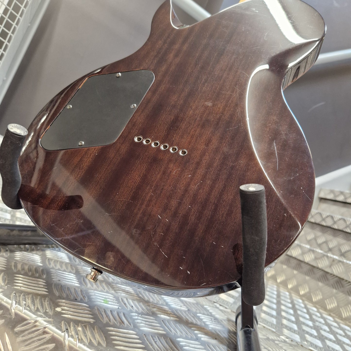 Pre-Owned Chapman ML-2 Electric - Grey Close-up of the back of a dark brown wooden electric guitar resting on a stand against a textured metal wall.