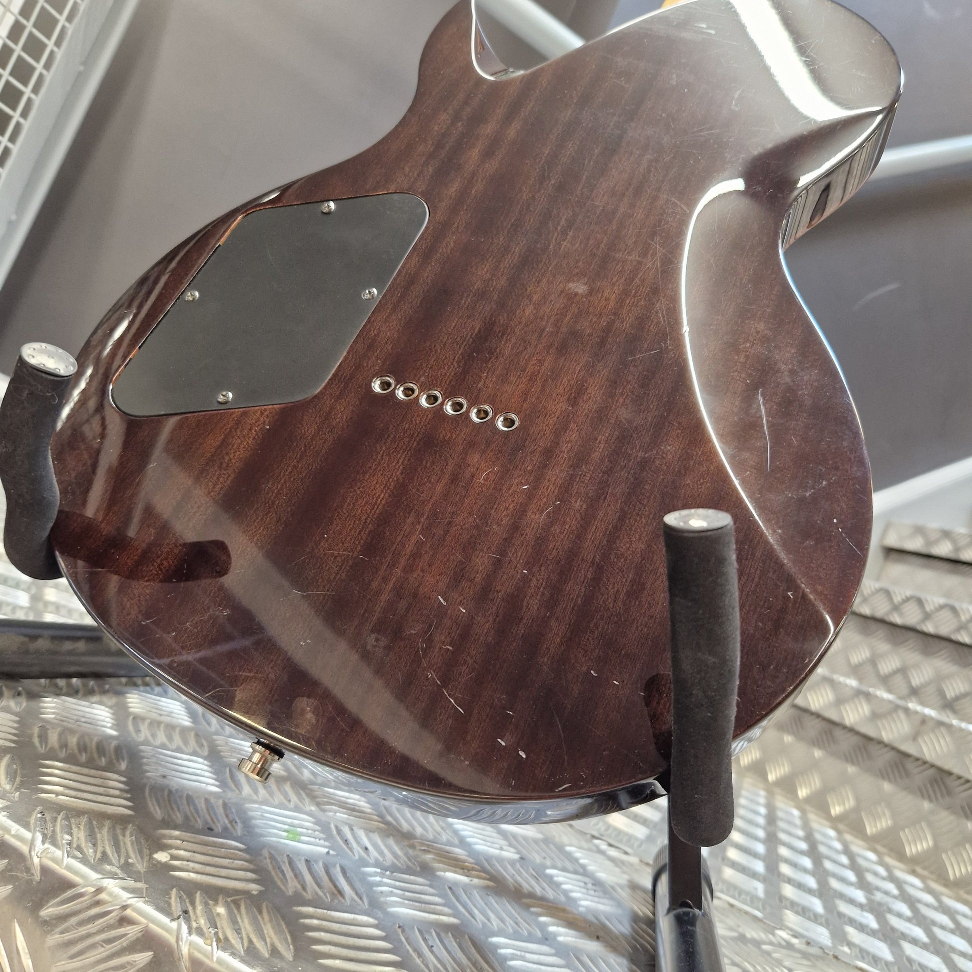 Pre-Owned Chapman ML-2 Electric - Grey Close-up of the back of a dark brown wooden electric guitar resting on a stand against a textured metal wall.