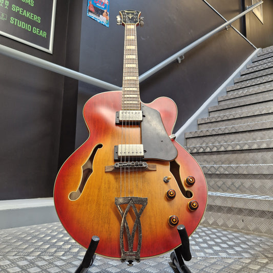 Pre-Owned Ibanez AFV75-VAL Artcore Hollowbody Electric - Vintage Amber Burst Low An electric guitar with a sunburst orange and brown body, mounted on a display stand in an indoor showroom setting.