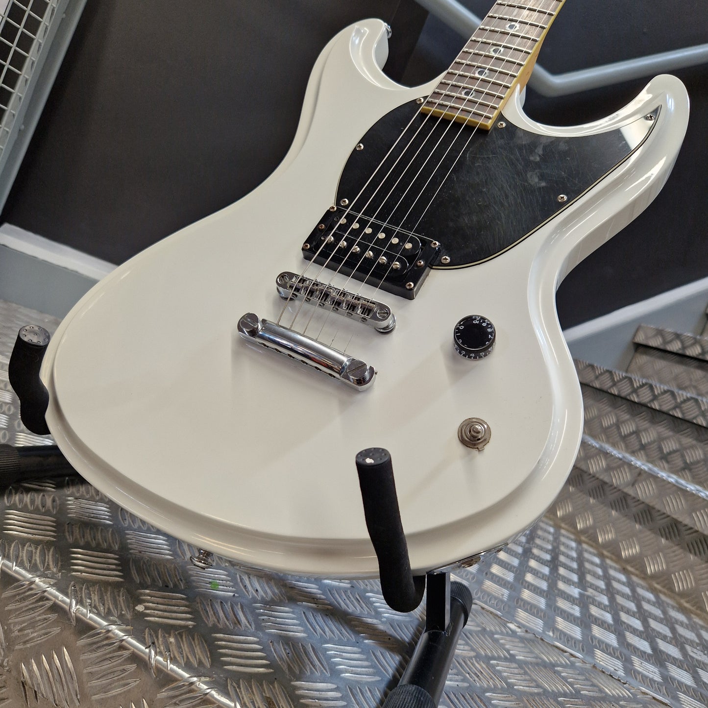 Pre-Owned Schecter Shaun Morgan Signature Electric - White A white electric guitar with a black pickguard and chrome hardware, photographed on a stand against a textured metal diamond plate background.