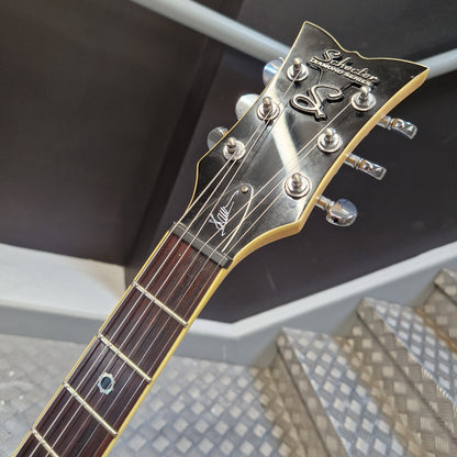 Pre-Owned Schecter Shaun Morgan Signature Electric - White Close-up of a guitar headstock and upper fretboard, showing a dark brown wood neck, gold-toned fretboard edges, silver tuning pegs, and a black headstock with white script branding.