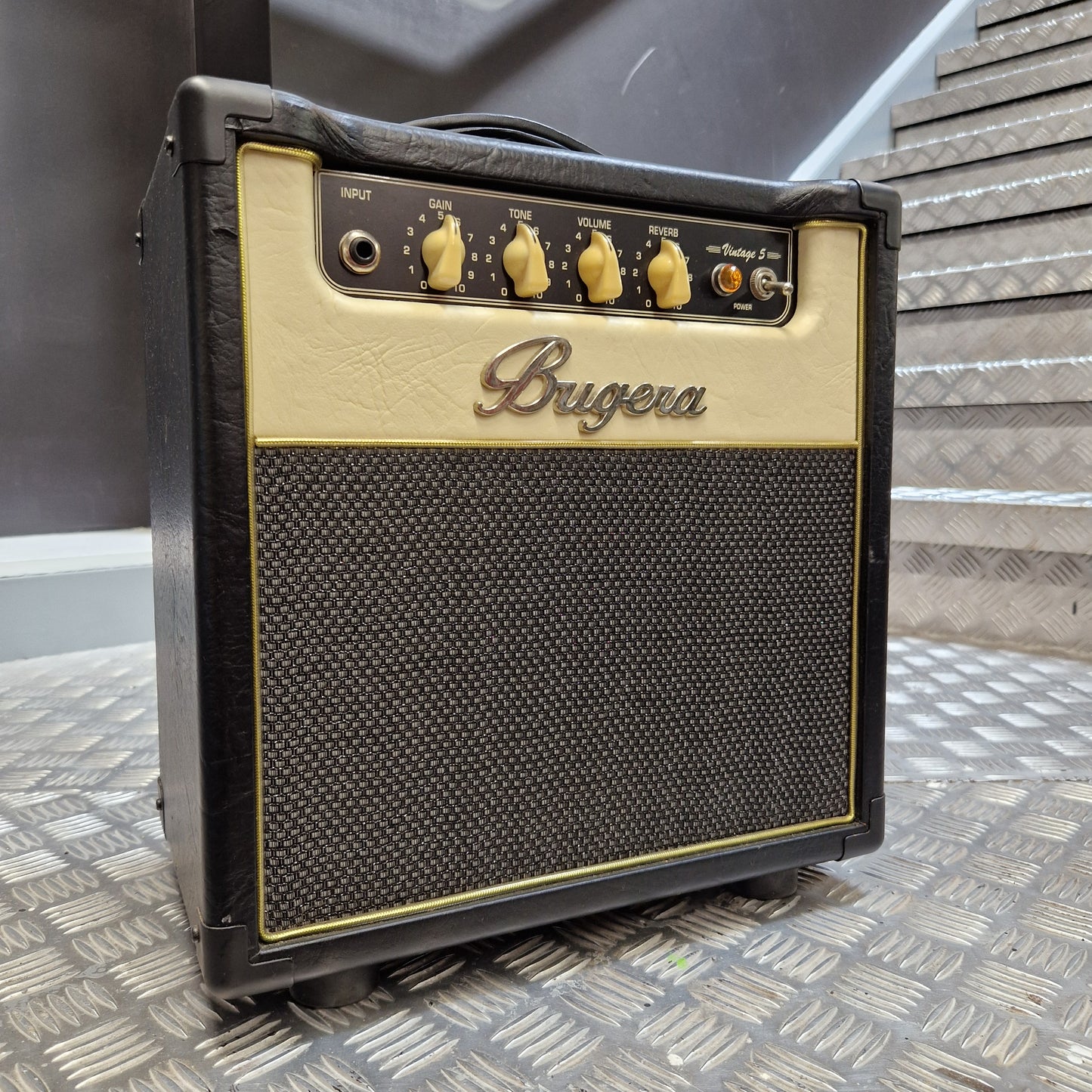 Pre-Owned Bugera Vintage 5 V5 Valve Guitar Combo A Bugga guitar amplifier head photographed from above, resting on a dark surface against a textured diamond-plate metal wall. The amp has a black body, a cream-colored faceplate, and gold controls and trim.
