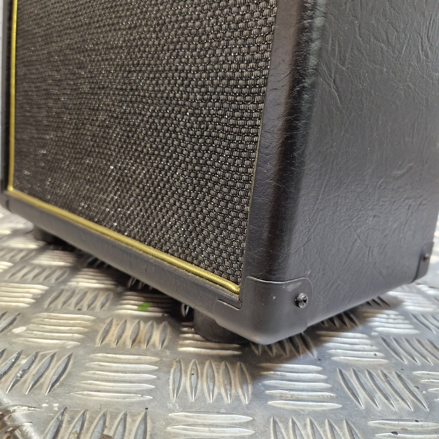 Pre-Owned Bugera Vintage 5 V5 Valve Guitar Combo A close-up of a black guitar amplifier or speaker cabinet corner with a textured grille and a thin yellow edge trim, resting on a silver diamond plate metal surface.