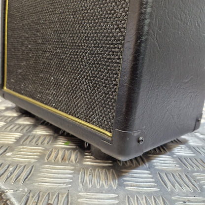 Pre-Owned Bugera Vintage 5 V5 Valve Guitar Combo A close-up of a black guitar amplifier or speaker cabinet corner with a textured grille and a thin yellow edge trim, resting on a silver diamond plate metal surface.
