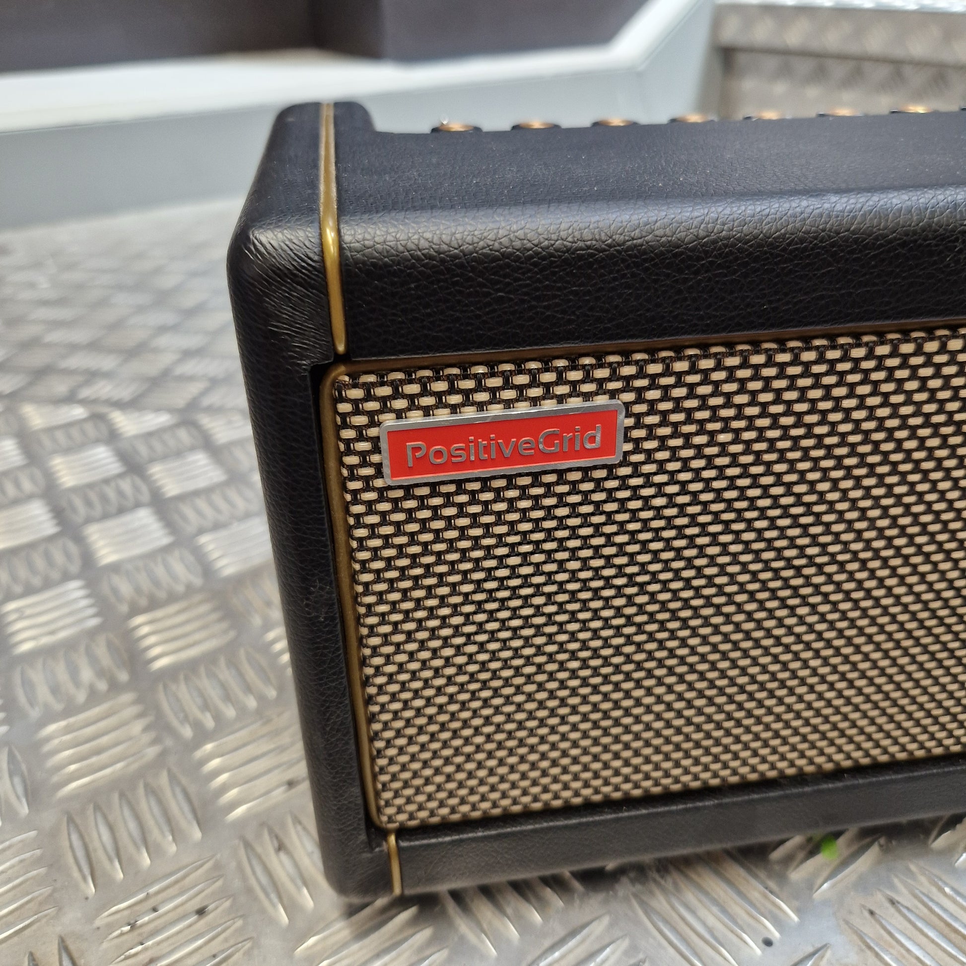 Pre-Owned Positive Grid Spark 40-Watt Smart Guitar Combo & Bluetooth Speaker A close-up of a Positive Grid speaker showing its black textured leather-like side, beige mesh grille, and a red brand badge, resting on a diamond plate metal surface.