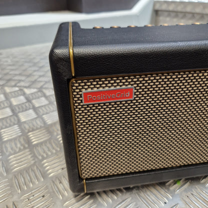 Pre-Owned Positive Grid Spark 40-Watt Smart Guitar Combo & Bluetooth Speaker A close-up of a Positive Grid speaker showing its black textured leather-like side, beige mesh grille, and a red brand badge, resting on a diamond plate metal surface.