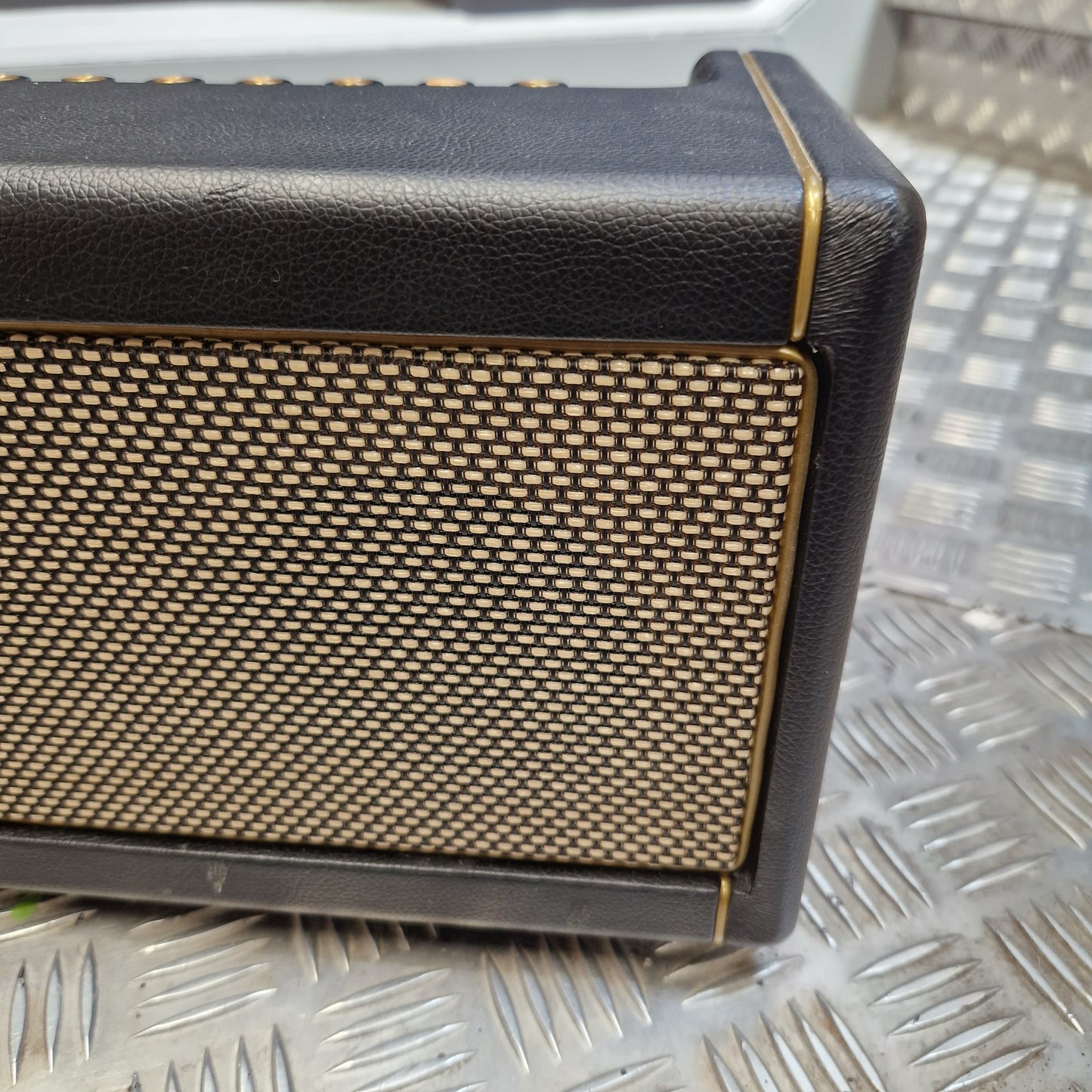 Pre-Owned Positive Grid Spark 40-Watt Smart Guitar Combo & Bluetooth Speaker A close-up of a black leather-look speaker or amplifier front panel with a gold mesh grille and thin gold trim lines, set against a textured silver metal background.