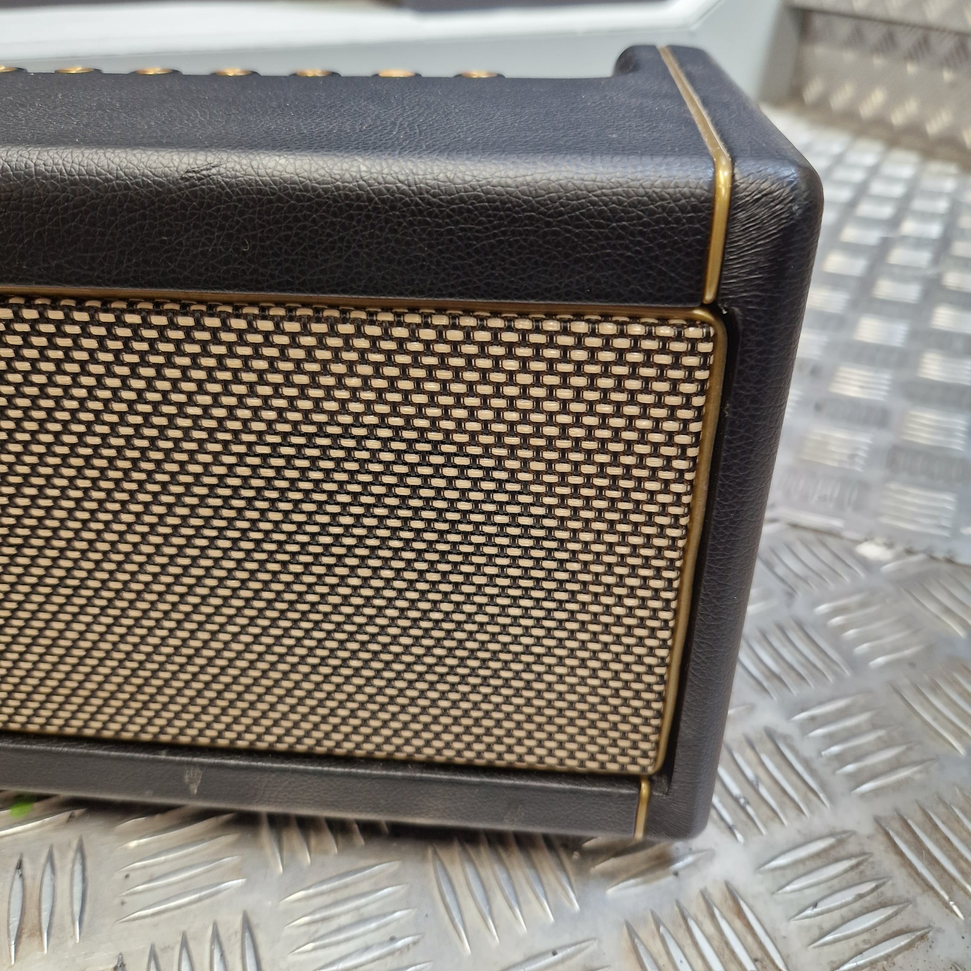 Pre-Owned Positive Grid Spark 40-Watt Smart Guitar Combo & Bluetooth Speaker A close-up of a black leather-look speaker or amplifier front panel with a gold mesh grille and thin gold trim lines, set against a textured silver metal background.