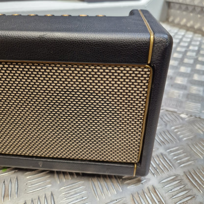 Pre-Owned Positive Grid Spark 40-Watt Smart Guitar Combo & Bluetooth Speaker A close-up of a black leather-look speaker or amplifier front panel with a gold mesh grille and thin gold trim lines, set against a textured silver metal background.
