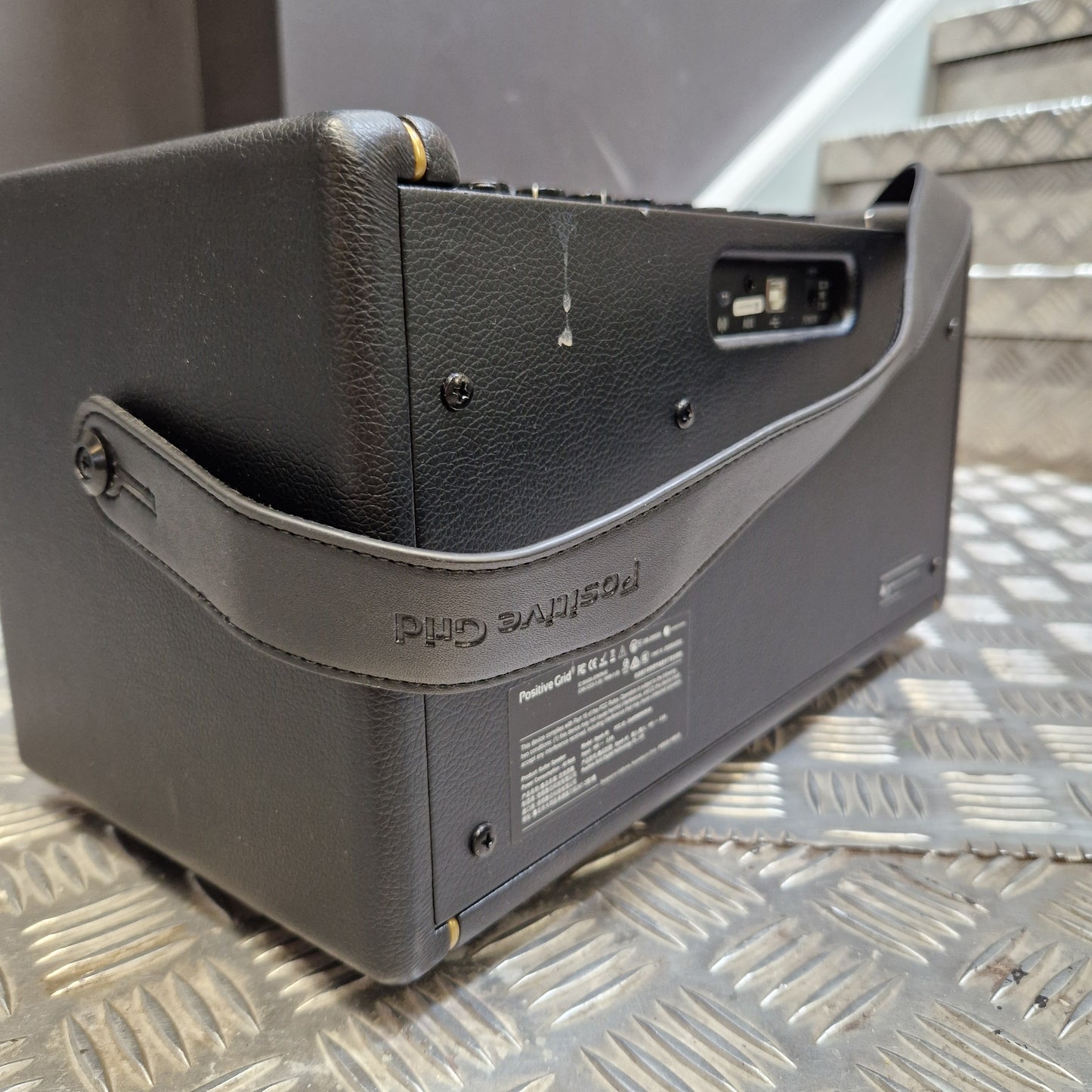 Pre-Owned Positive Grid Spark 40-Watt Smart Guitar Combo & Bluetooth Speaker A black textured guitar amplifier or speaker cabinet with a matching shoulder strap, photographed on a diamond-plate metal surface.
