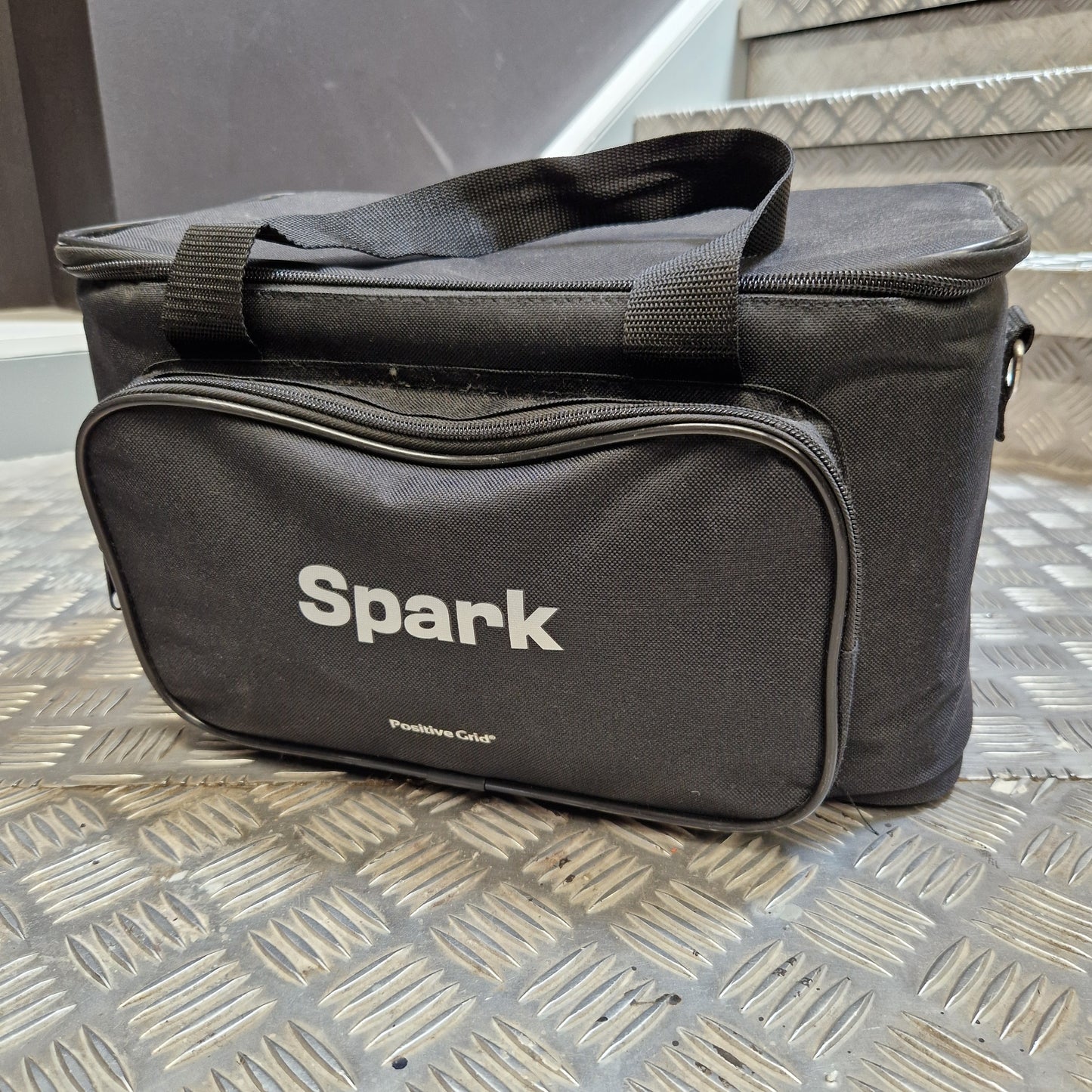 Pre-Owned Positive Grid Spark 40-Watt Smart Guitar Combo & Bluetooth Speaker A black fabric bag with a white 'Spark' logo printed on the front pocket, resting on a diamond plate metal surface.