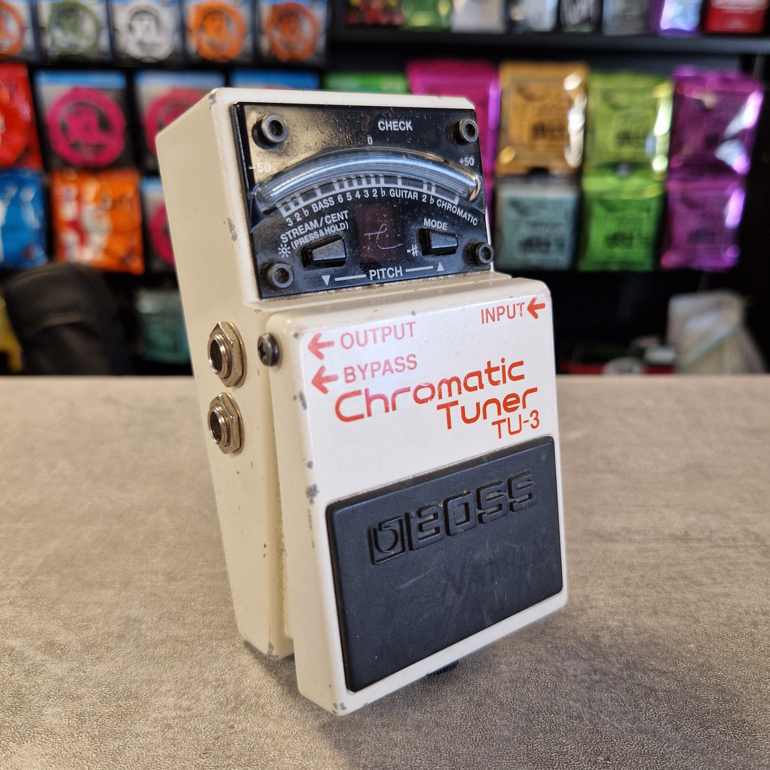 Pre-Owned Boss TU-3 Tuner Pedal A Boss Chromatic Tuner TU-3 guitar pedal with a cream-colored metal casing, a black footswitch, and a black dials faceplate, shown on a beige surface with a blurred background of colorful items.