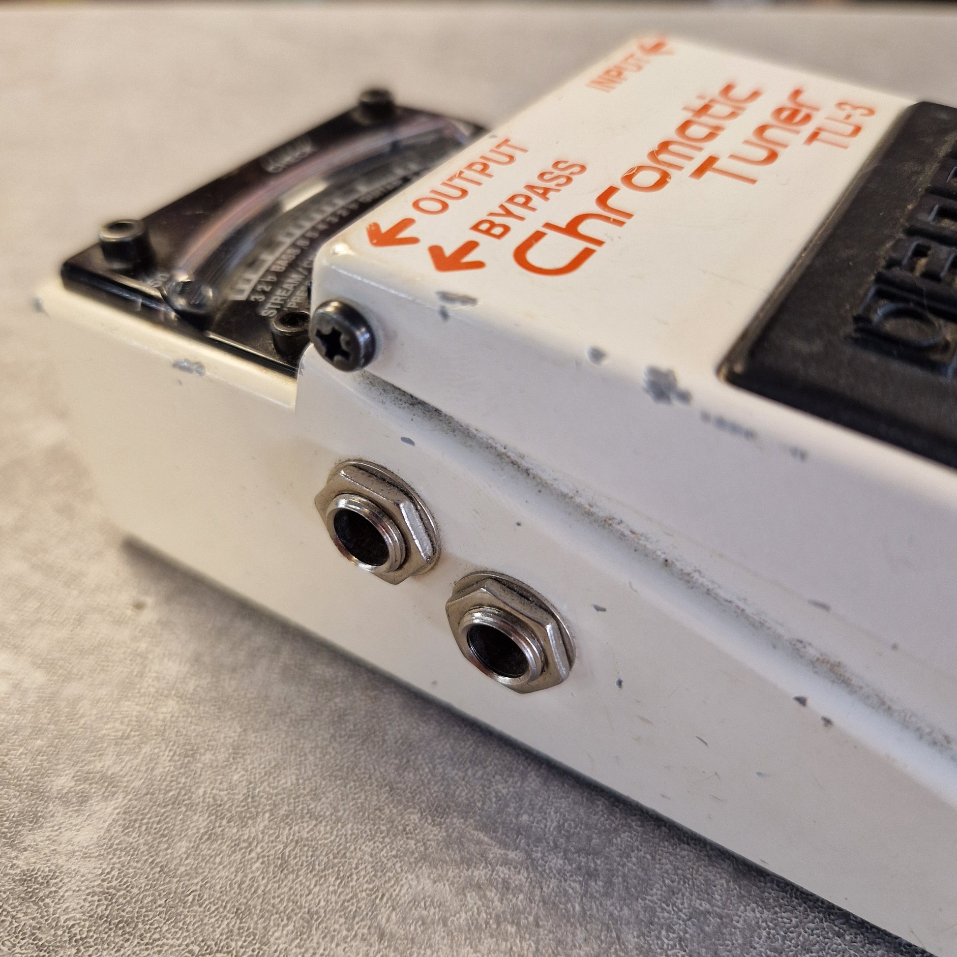Pre-Owned Boss TU-3 Tuner Pedal Close-up of a white Chromatic Tuner TU3 guitar pedal showing the orange printed branding, output and bypass jacks, and part of the black volume control on a gray textured surface.