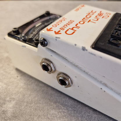 Pre-Owned Boss TU-3 Tuner Pedal Close-up of a white Chromatic Tuner TU3 guitar pedal showing the orange printed branding, output and bypass jacks, and part of the black volume control on a gray textured surface.