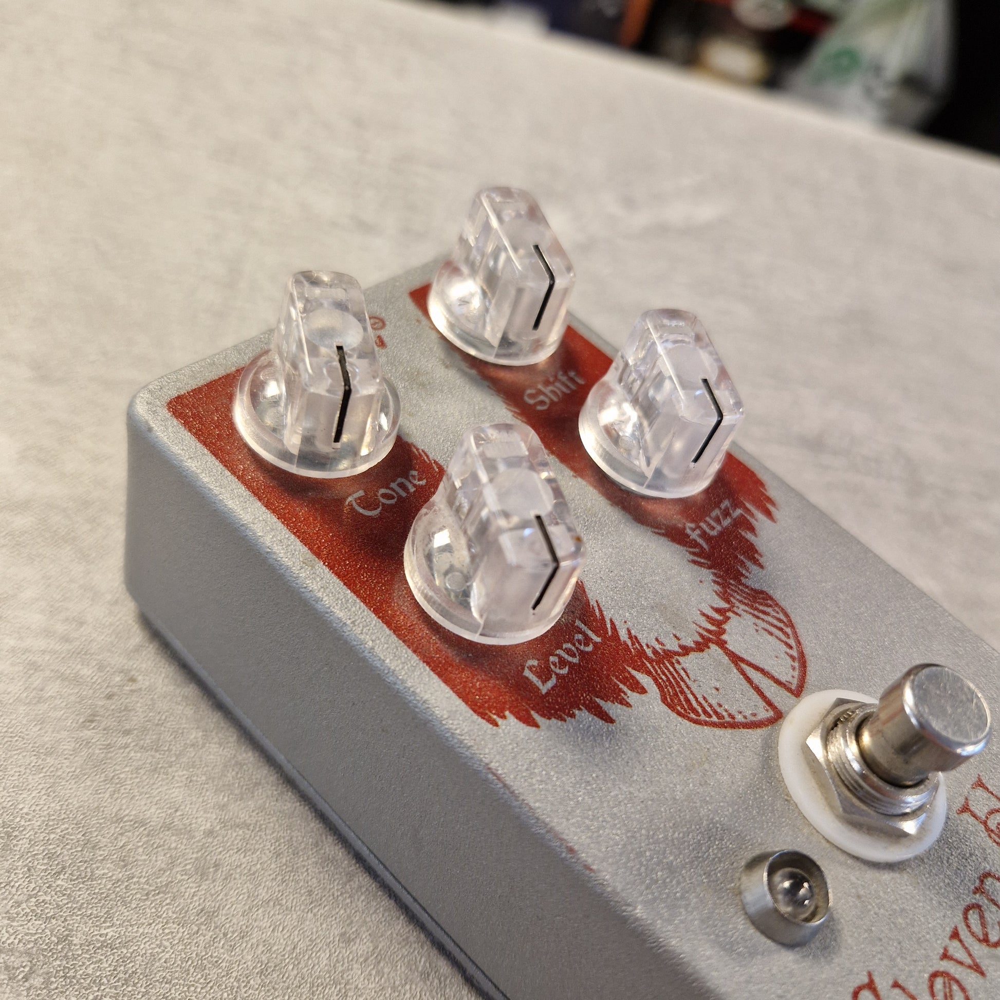 Pre-Owned EarthQuaker Devices Cloven Hoof Fuzz Pedal A close-up of a silver guitar effects pedal with four clear plastic knobs, red printed graphics, and a metal footswitch on a gray textured surface.