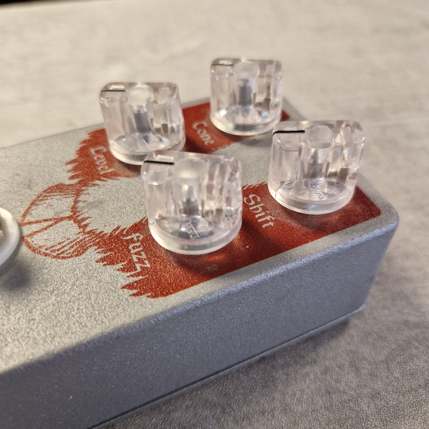 Pre-Owned EarthQuaker Devices Cloven Hoof Fuzz Pedal A close-up of a guitar effects pedal with a textured gray body, red printed graphics, and four clear acrylic knobs labeled Gain, FX1, FX2, and Com.