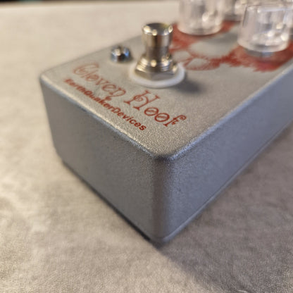 Pre-Owned EarthQuaker Devices Cloven Hoof Fuzz Pedal A close-up shot of a Worldbucker Devices Clever Kloof guitar effects pedal, showing its textured gray metal casing, footswitch, and clear knobs on a neutral gray surface.