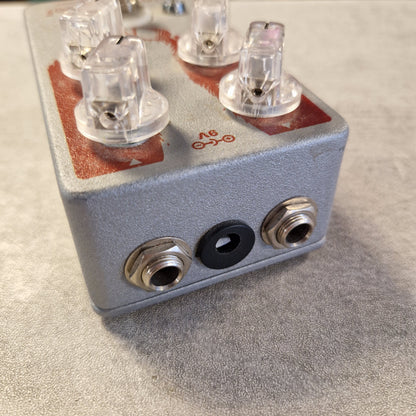 Pre-Owned EarthQuaker Devices Cloven Hoof Fuzz Pedal A close-up of a small metal electronic component with clear plastic connectors and two silver jack-style ports on a textured gray surface.