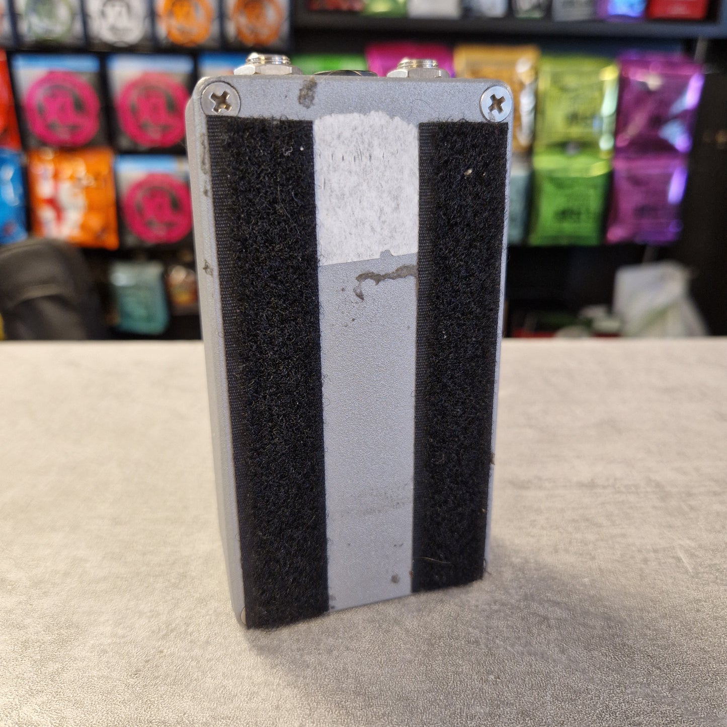 Pre-Owned EarthQuaker Devices Cloven Hoof Fuzz Pedal A rectangular metal device with black textured side panels and a light gray center section, standing upright on a tabletop with a blurred store background.