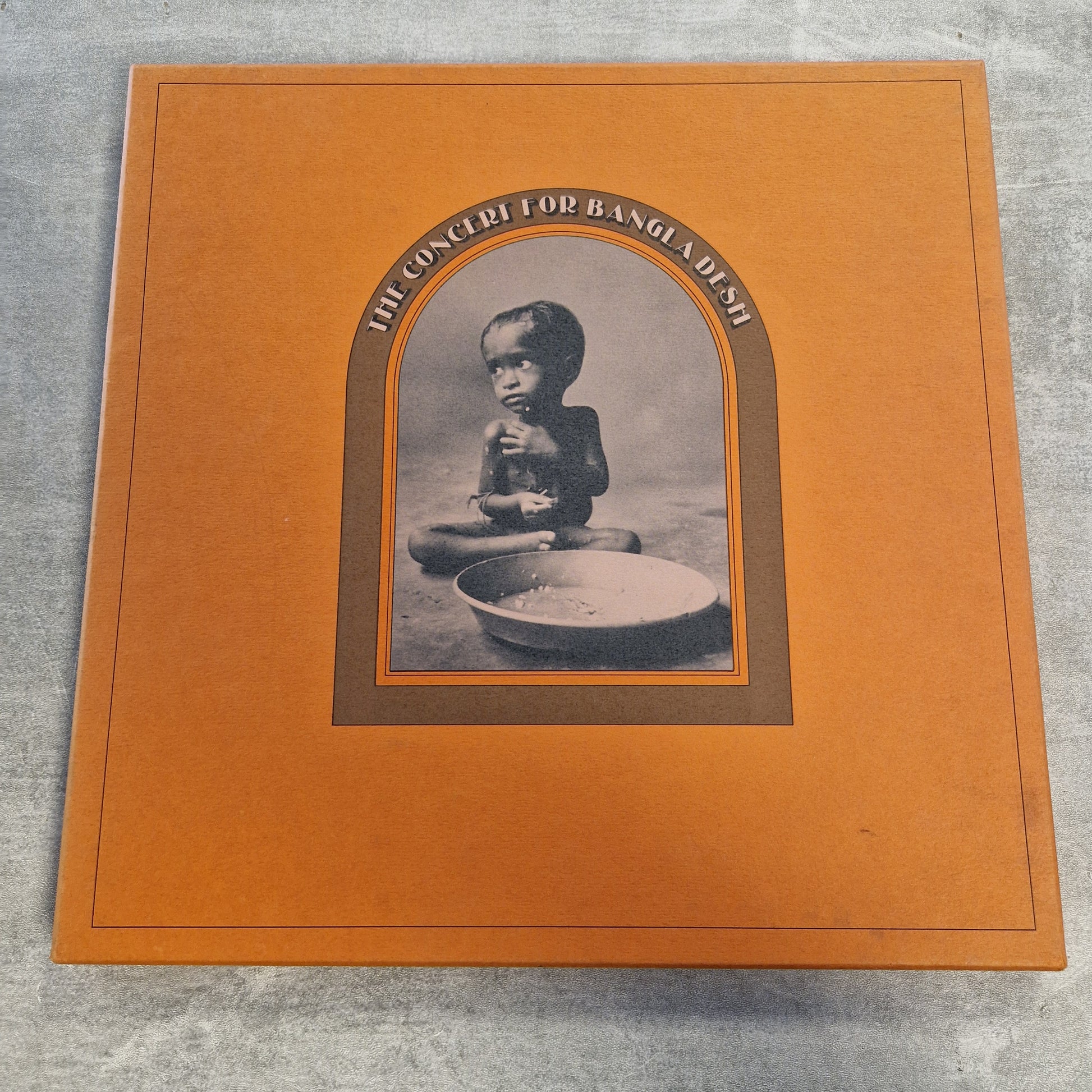 Various - The Concert For Bangla Desh Vintage Vinyl Boxset A square orange album cover or book-style package photographed on a gray textured surface. The center features an arched inset with a black-and-white photo of a child and the text 'HE'S GOT TO BE A MAD-ASS' around the image.