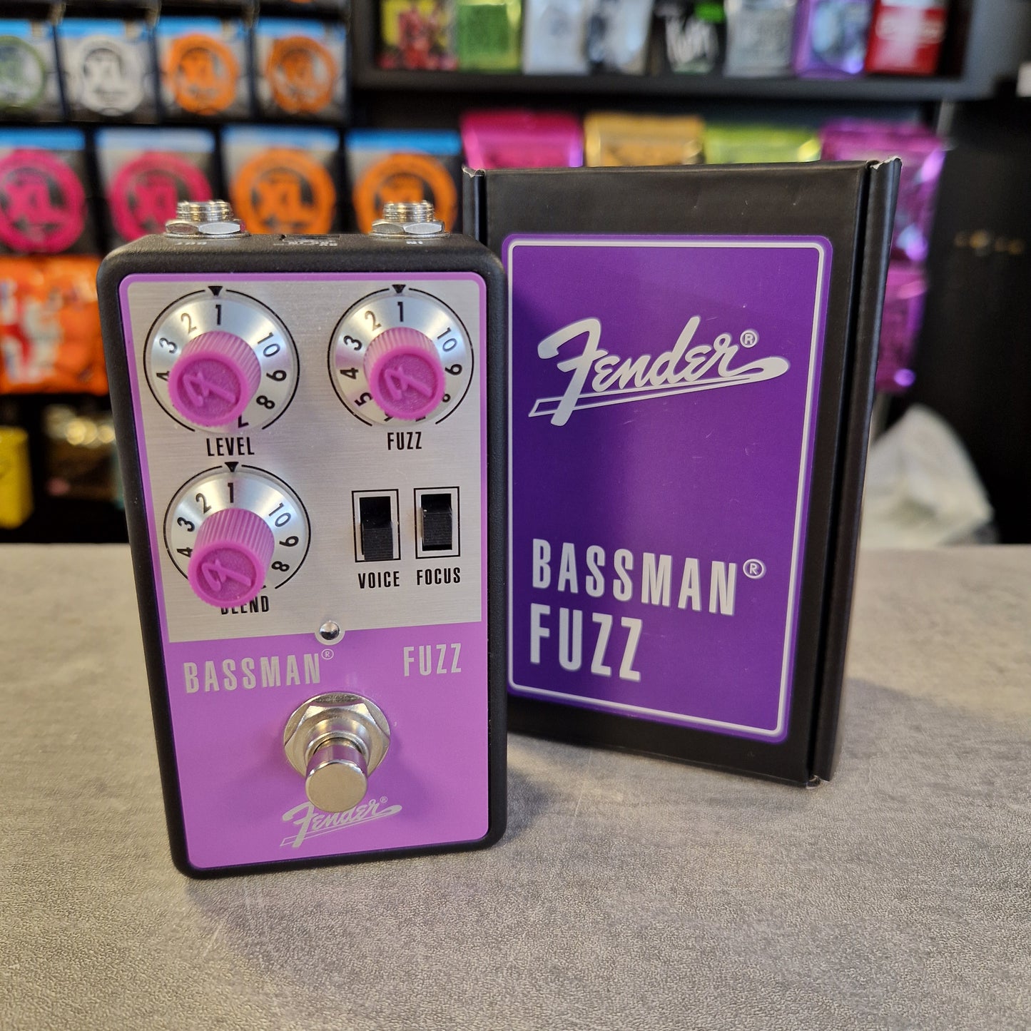Pre-Owned Fender Bassman Fuzz Pedal A Fender Bassman Fuzz guitar effects pedal with a purple faceplate and silver controls, standing next to its black and purple retail box on a gray surface. The background shows a blurred store shelf with colorful items.