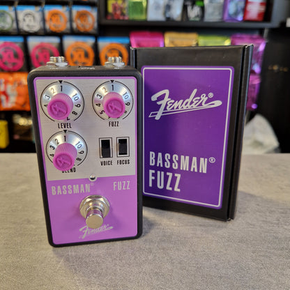 Pre-Owned Fender Bassman Fuzz Pedal A Fender Bassman Fuzz guitar effects pedal with a purple faceplate and silver controls, standing next to its black and purple retail box on a gray surface. The background shows a blurred store shelf with colorful items.