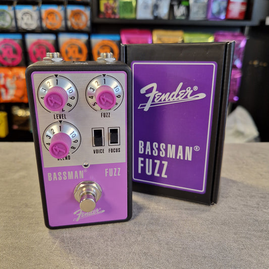 Pre-Owned Fender Bassman Fuzz Pedal A Fender Bassman Fuzz guitar effects pedal with a purple faceplate and silver controls, standing next to its black and purple retail box on a gray surface. The background shows a blurred store shelf with colorful items.